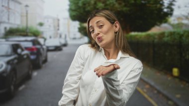 Beyaz gömlekli, katlanmış kollarıyla gülümseyen genç sarışın kadın park etmiş arabaları ve yapraklı çiti olan bir şehir caddesinde, düzgün görünümlü, gündelik sıcak neşe.