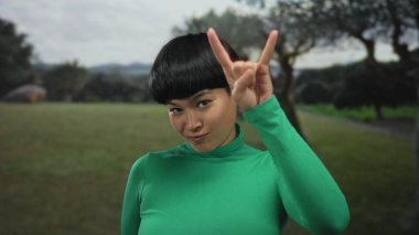Yeşil elbiseli genç bir kadın dışarıda, yeşil bir parkta, neşeli bir ifadeyle rock 'n roll hareketi yapıyor..
