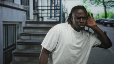 Kulak kabartan ve caddede öne doğru eğilen adam beton adımlar ve park edilmiş arabalar; meraklı dinleme niyeti.