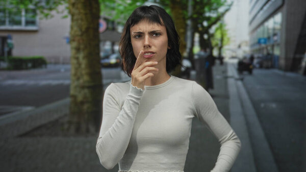 Young hispanic woman pinches nose with hand raised on busy street under leafy trees near concrete sidewalk and modern buildings; disgust.