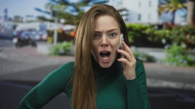 Binalar ve araçlarla dolu kalabalık bir şehir sokağında akıllı telefonu kulağına dayayan kadın; öfke.