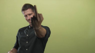 Kireç yeşili stüdyoda orta parmak ve çıplak el bileziği takan adam dik dik bakıyor ve kollarını kaldırıyor..