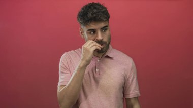 Kırmızı stüdyo ortamında tırnak yiyen ve yan yan bakarken baş parmağıyla alt dudağa bastıran bir adam..