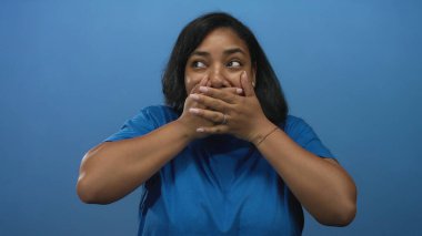 Mavi tişört giyen Afro-Amerikalı kadın elleriyle ağzını kapatıyor. Stüdyoda mavi arka planda çıplak kollar ve nazik bir gülümseme sergiliyor..