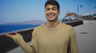 Bir adam deniz kenarında yürüyüş yaparken elini kaldırıyor. İspanyol bir adam açık hava manzarasından hoşlanıyor. Karşılama jesti yapıyor ve gülümsüyor..