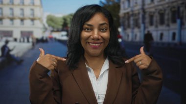 Kadın gülerek ve parmakla göğsünü göstererek şehir caddesinde gün ışığında kahverengi ceket giyiyor. Kendine güven..