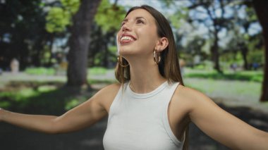 Altın halkalı küpeleri olan beyaz tişörtlü bir kadın kollarını parkta çıplak omuzlarıyla açar..