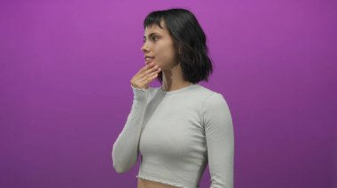 Açık gri gömlekli genç İspanyol kadın mor stüdyoda hafif kaşları ve çilleri olan çeneye dokunur..