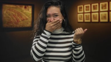 Bir sanat galerisi içinde kadın eli ağızla kaplıdır, başparmağını geriye uzatır ve çizgili balıkçı yaka kazak ve tahta saat giyerken güler; sürpriz zevk.