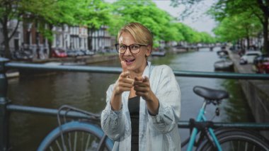 Kanal caddesinde park halindeki bisikletin yanındaki kamerayı işaret eden kadın, gözlük ve açık renk kot gömlek giyiyor, gülümsüyor, kendine güveni tam..