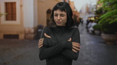 Siyah spor kıyafetli genç İspanyol kadın kış şafağında kaldırım taşı sokağında soğuk ellerini ovuşturuyor. Kararlılık..