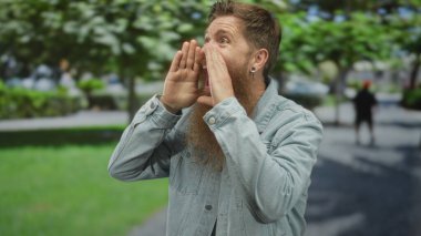 Uzun sakallı bir adam orman ortamında elleri ağızlarına sıkışmış bir şekilde bağırarak kot ceket giyiyor..