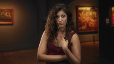 Kadın, çerçeveli tablolar ve sıcak yukarıdan aydınlatma arasında bir sanat galerisi içinde kolları çaprazlanmış bir izleyiciyi işaret ediyor; şüphecilik iddiası.