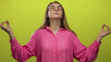 Pembe gömlekli bir kadın, izole edilmiş sarı arka plana karşı meditasyon yapıyor. Yoga Mudra 'da elleri ile sakinliğini ve rahatlığını ifade ederken günlük hayatın stresine karşı koyuyor..