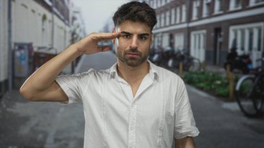 Latin kökenli genç adam, sokağın köşesinde, yumuşak güneş ışığı altında gündelik selamlama jesti ile elini kaldırır; sıcak selamlaşma.