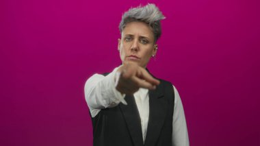 Kısa gri saçlı, siyah yelek giyen ve beyaz gömlekli bir kadın doğrudan bakışlı, eflatun renkli stüdyoyu işaret ediyor..