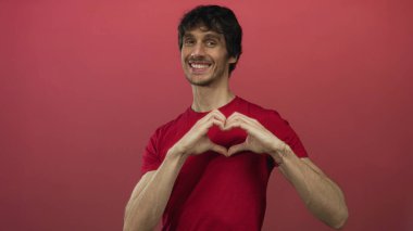 Kırmızı gömlekli genç adam gülümsüyor ve elleri stüdyoda kırmızı arkaplanda kalp hareketi yapıyor..