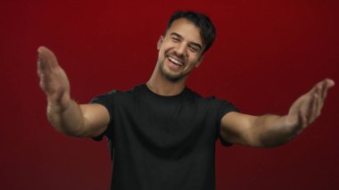Siyah tişörtlü genç İspanyol adam kendini izole edilmiş kırmızı arka planda kucaklarken sıcak bir şekilde gülümsüyor..