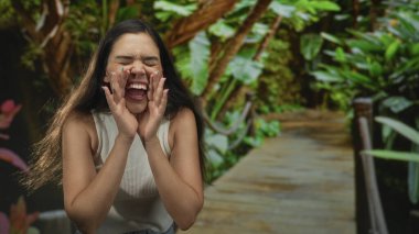 Yeşil yeşillikler ve bitkilerle çevrili orman parkında güneşli bir tahta kaldırımda elleri ağızda bağıran genç bir kadın..
