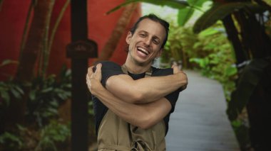 Bir orman geçidinde çıplak kollarını saran adam yemyeşil tropikal bitkiler ve ahşap bir levha direğiyle kaplı, kapalı gözlerle gülümseyen; kişisel bakım sevinci.