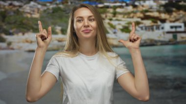 Arka planda bulanık deniz manzaralı, manzaralı bir plajda mutlu bir şekilde el kol hareketi yapan genç kadın rahat açık hava keyfi ve pozitifliğini sergiliyor..