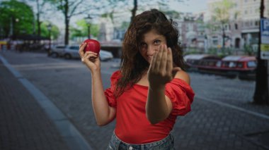Kadın kırmızı bir üst gülümseme içinde şehir caddesinde uzanmış elleri ile kırmızı elma tutar ve çağırır; neşeli bir davet.