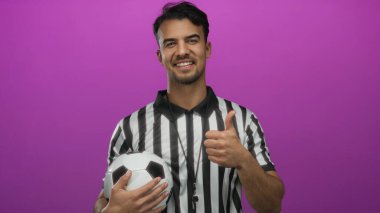 Hakem üniforması giyen İspanyol genç adam, pembe arka planda futbol topunu tutuyor, pozitifliğini ve spor heyecanını dile getiriyor..