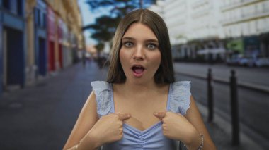 Woman pointing finger to chest on a crowded urban street in front of historic buildings with uncertain expression; confusion.