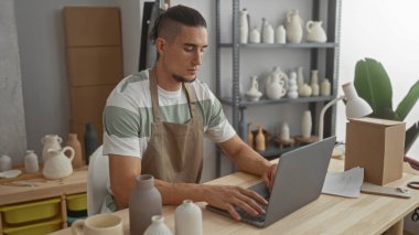 Stüdyo çalışma alanında çömlek vazoları ve raflarla çevrili ahşap masadaki klavyede önlük ve ellerle dizüstü bilgisayarda yazan adam; sakin yaratıcılığa odaklan.