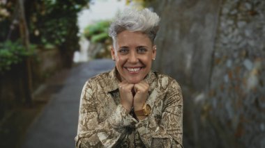 Kısa saçlı bir kadın, taş duvarlı ve yeşillikli dar bir sokakta yumruklarını sıkıyor. Sürpriz..