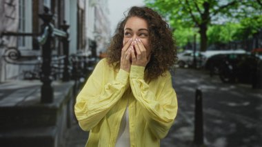 Sarı tişörtlü ve beyaz tişörtlü kadın şehir caddesinde, kıvırcık saçlı ve görünür sırıtışlı el hareketi yaparken; şehir neşesi.
