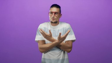 Kırmızı gözlüklü ve çizgili tişörtlü bir adam stüdyoda kameraya yan gözle bakarken kollarını dışa doğru çeviriyor..