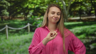 Pembe tişörtlü genç bir kadın yeşil bir parkta duruyor, yüzünde bir gülümsemeyle, parlak doğal ışık altında, açık havada etkileyici vücut dilini gösteriyor..