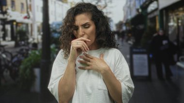 Beyaz gömlekli kadın, gün ışığında hareketli şehir caddesinde ayakta dururken ağzını eliyle ve öksürüğüyle kapatıyor..