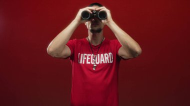Kırmızı cankurtaran tişörtü giyen ve boynunda ıslık çalan bir adam iki eliyle siyah dürbünü tutuyor ve ön kollarını stüdyonun kırmızı arka planına dayıyor..