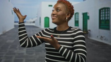 Çizgili kazaklı kadın, güneşli caddede beyaz binalarla kaplı avuçlarıyla ellerini sunar; sıcak karşılama.