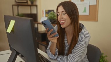 Ağzı açık bir kadın akıllı telefona doğru eğilir ve dağınık ofis masasında bilgisayar monitörü ve mantar panoya yapıştırılmış yapışkan notlarla mavi telefona konuşur;.