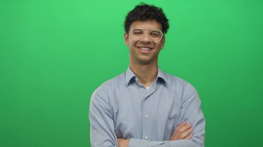 Genç İspanyol gözlüklü adam kollarını kavuşturuyor ve iyi aydınlatılmış bir stüdyoda sıcak bir gülümseme sergiliyor..