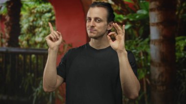 Siyah tişörtlü adam, hafif bir sırıtışla gülümserken el ele işaret ediyor ve işaret veriyor..