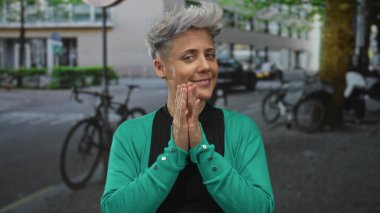 Kadın, yapraklı ağaçların altında bisiklet ve park halindeki arabalarla dolu dar bir kaldırım taşı sokakta el çırpıyor..