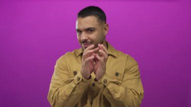 Kısa koyu renk saçlı, kahverengi ceketli, el çırpan bir adam parlak mor stüdyo duvarına bakarken, huzur içinde..