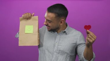 İnsan, stüdyoda küçük kırmızı kalp şeklindeki kahverengi kağıt torbayı ve yapışkan gülücük notunu tutar; mutluluk sevgisi sıcaklığı.