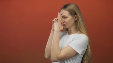 Kafkasyalı genç bir kadın, izole edilmiş kırmızı arka plan duvarındaki silaha benzeyen bir el hareketi yapıyor, beyaz bir tişört giyiyor, ciddi bir ifadeyle oyunculuğu çağrıştırıyor..