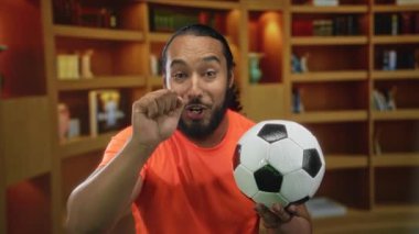 Kitap rafları arasında kütüphane binasında gülümserken futbol topu tutan ve yumruk kaldıran turuncu gömlekli adam; takım ruhu heyecanı.