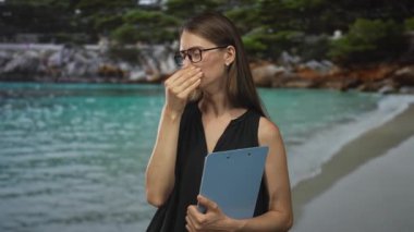 Gözlüklü bir kadın elinde mavi bir pano ve bir eli kumlu bir sahil kıyısında güneşe bakarken alnına bastırılmış bir şekilde çalışıyor..