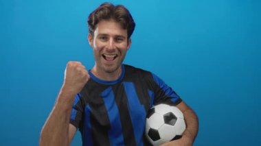 Futbol topu tutan, yumruğunu sıkan ve mavi stüdyoda siyah ve mavi forma giyerken gülümseyen bir adam..