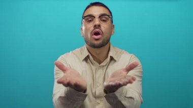 Latin asıllı genç adam bej gömlekli, öpüşen ve elleri açık bir şekilde Teal stüdyosu ortamında kameraya doğru eğiliyor..