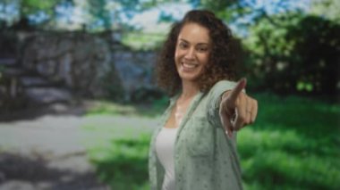 Kadın, uzanan koluyla gülümserken taş duvarın ve yeşil çimlerin yanındaki ormanda kamerayı işaret ediyor; keyif daveti.