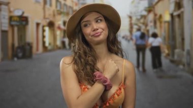 Turuncu bikinili ve geniş hasır şapkalı kadın, güneş ışığı altındaki bir kaldırım taşı caddesinde sessizlik için parmaklarını dudaklarına koyar..