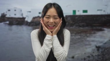 Genç Çinli kadın deniz kenarında mutlu bir şekilde gülümsüyor. Arka planda beyaz binalar, sakin bir sahil gününde..